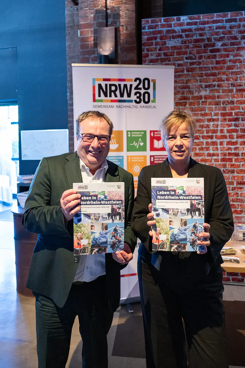 Ein Mann und eine Frau halten die neue Nachhaltigkeitsstrategie.NRW 2026 „Leben in Nordrhein-Westfalen“ vor dem NRW-2030-Aufsteller in die Kamera