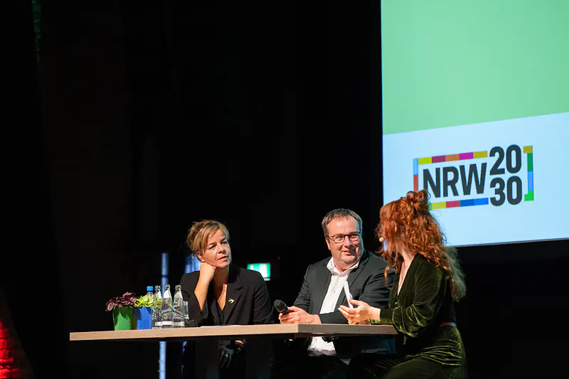 Bühnengespräch zur Nachhaltigkeitsstrategie.NRW 2026: zwei Gäste und die Moderatorin am Tisch vor dem NRW-2030-Logo