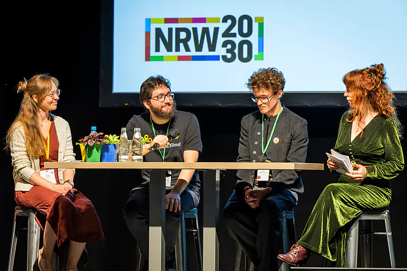 Drei junge Gäste im Bühnengespräch mit der Moderatorin vor dem NRW-2030-Logo