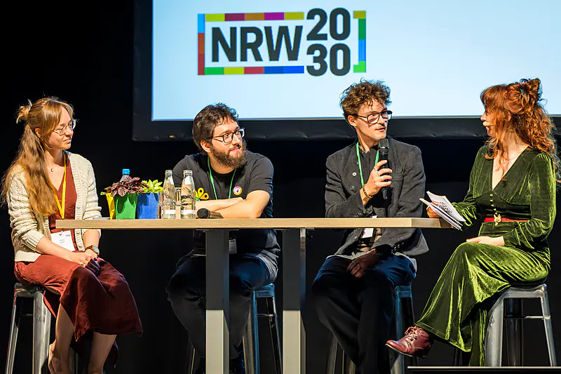 Ein junger Gast spricht in der Talkrunde ins Mikrofon, die Moderatorin hört mit ihren Moderationskarten zu
