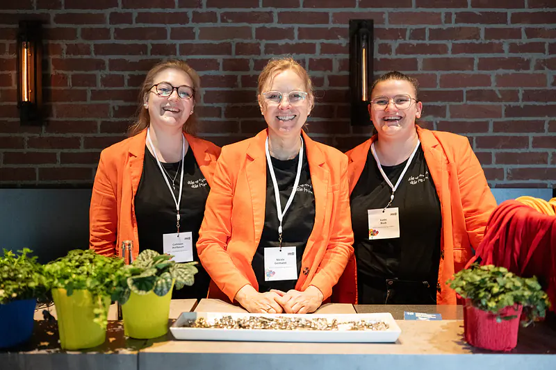 Drei Frauen aus dem Team in orangefarbenen Blazern lachen am Empfangstresen